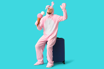 Wall Mural - Young man in bunny costume with ice-cream and suitcase on blue background. Easter celebration