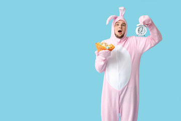 Canvas Print - Shocked young man in bunny costume holding wicker basket with Easter eggs and alarm clock on blue background