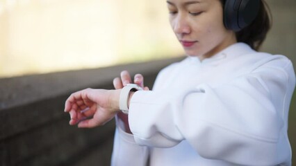 Wall Mural - Asian woman listening to music with headphones and smart watch mobile app during jogging exercise at city street in the morning. Healthy girl do sport training running workout in the city.