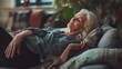Cheerful sleepy blonde older lady lying on couch, leaning legs and feet on soft back, enjoying leisure, lazy break in modern home interior, breathing fresh cool pure air with closed eyes.