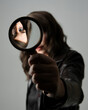 © faestock - close up portrait of beautiful brunette woman wearing black leather trench coat. Holding detective magnifying glass, reaching out towards camera, searching for discovery. Isolated  studio background