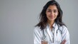 Portrait of an attractive Indian female physician wearing a white lab coat and stethoscope, with a friendly expression on a solid gray backdrop.