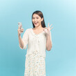 Beautiful young asian woman in white dress with flower pattern using smartphone show okay sign isolated on blue background