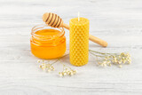 Fototapeta Sypialnia - Open glass jar with honey, beeswax candle, honey dipper and white flower on a light wooden background, front view, selective focus. Alternative medicine, healthy eating.