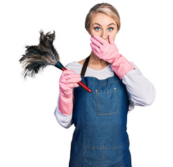 Canvas Print - Beautiful young blonde woman wearing apron holding cleaning duster shocked covering mouth with hands for mistake. secret concept.