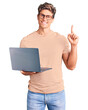 © Krakenimages.com - Young handsome man wearing glasses holding laptop surprised with an idea or question pointing finger with happy face, number one