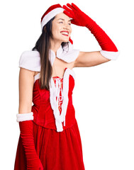 Sticker - Young beautiful caucasian woman wearing santa claus costume very happy and smiling looking far away with hand over head. searching concept.