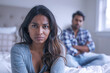 Indian woman sad in bedroom, her husband is leaning on the bed