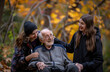 © Kien - An elderly man in his wheelchair is surrounded by two young women, one of them has her hand on the backrest and another woman stands behind him smiling at each other