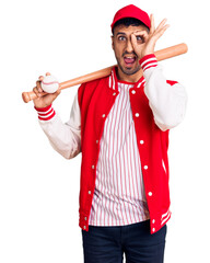 Wall Mural - Young hispanic man playing baseball holding bat and ball smiling happy doing ok sign with hand on eye looking through fingers