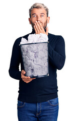 Poster - Young handsome blond man holding paper bin with crumpled papers covering mouth with hand, shocked and afraid for mistake. surprised expression