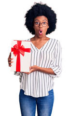 Wall Mural - Young african american woman holding gift scared and amazed with open mouth for surprise, disbelief face