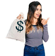 Wall Mural - Beautiful hispanic woman holding dollars bag pointing thumb up to the side smiling happy with open mouth