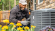 © evastar - HVAC Maintenance in a Blooming Garden. A technician inspects and repairs an outdoor HVAC unit surrounded by vibrant yellow flowers, with a house in the background. professional maintenance service in