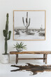 A white wall with a wood frame and cactus print, rustic wooden bench in front of it, white floor with cowhide rug, black and white photo of desert landscape, green plants on the side
