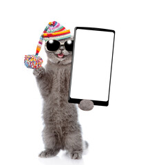 Aufkleber - Happy cat wearing warm knitted hat with pompon and sunglasses holding smartphone with white blank screen in it paw. Isolated on white background