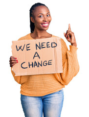 Wall Mural - Young african american woman holding we need a change banner surprised with an idea or question pointing finger with happy face, number one