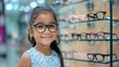 A young Mexican girl wearing glasses smiling as the child kid stands in front of a display case of glasses at an eye retail store