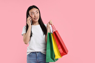 Wall Mural - Smiling woman with shopping bags talking by smartphone on pink background. Space for text