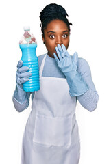 Wall Mural - Young african american woman wearing apron holding detergent bottle covering mouth with hand, shocked and afraid for mistake. surprised expression