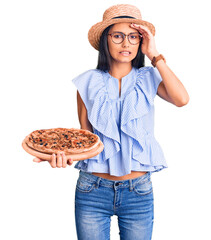 Canvas Print - Young beautiful latin girl wearing summer hat and glasses holding pizza stressed and frustrated with hand on head, surprised and angry face