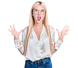 Wall Mural - Beautiful blonde woman wearing elegant shirt and glasses crazy and mad shouting and yelling with aggressive expression and arms raised. frustration concept.