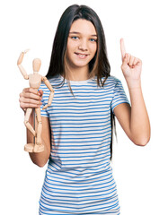 Poster - Young brunette girl with long hair holding small wooden manikin smiling with an idea or question pointing finger with happy face, number one