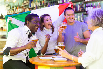 Wall Mural - Extremely happy italian man and woman supporters waving national flag, celebrating victory with glasses of beer in beer pub