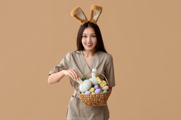 Poster - Female Asian massage therapist in bunny ears with Easter eggs and herbal bags on brown background