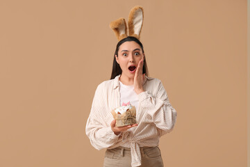 Canvas Print - Shocked young woman in bunny ears with sweet Easter cake on brown background