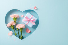 Mother's Day Sophisticated Display, From Top View, Featuring Carnations, A Gift Wrapped In Silk, Paper Hearts, All Visible Through Heart-shaped Hole On Pastel Blue Background, With Empty Space