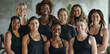 A group of diverse, happy women wearing black sportswear, standing together in a gym setting.