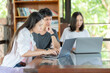 © Nutchapong Wuttisak - Side view of an Asian female student studying online on a laptop and two friends in the library. They are studying in a university group and read books every week to gain knowledge.