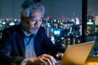 Portrait of mature Asian businessman working on laptop computer in office at night
