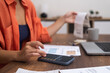 Expense Management: Seated at a desk with a laptop, a woman uses her smartphone calculator to compute household finances