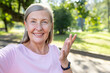Close-up photo of an active senior gray-haired woman walking in the park, talking on the phone with a video camera, smiling and gesturing with her hands.