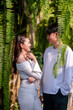 © Wasana - Young couple sharing a loving gaze among hanging ferns in a tranquil outdoor garden setting.