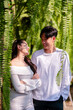 © Wasana - Young couple sharing a loving gaze among hanging ferns in a tranquil outdoor garden setting.