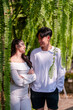 © Wasana - Young couple sharing a loving gaze among hanging ferns in a tranquil outdoor garden setting.