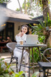 © Wasana - A happy young woman enjoys remote work on her laptop, comfortably seated outdoor of cafe with a coffee.
