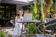 © Wasana - A young woman talking on the phone while working on a laptop in a quiet outdoor cafe.