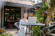 © Wasana - A happy young woman enjoys remote work on her laptop, comfortably seated outdoor of cafe with a coffee.