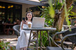 © Wasana - A young woman talking on the phone while working on a laptop in a quiet outdoor cafe.