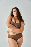 Fototapeta Na drzwi - alluring plus size young woman in underwear with curly brown hair hugging herself on grey background
