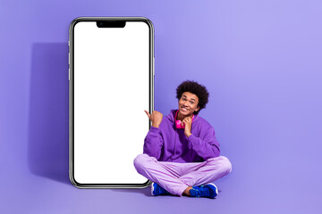 Sticker - Full length photo of optimistic nice guy sitting indicating at smartphone screen touching headphones isolated on violet color background