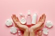 Woman's hands holding and presenting various skincare and cosmetic products against a pink background