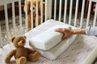 person placing an orthopedic pillow in a baby crib, teddy bear beside