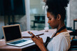 © sorapop - Close up portrait of focused black woman coffee shop owner in apron using smartphone in her cafe. Multi tasking barista manages orders communicates runs her own business.
