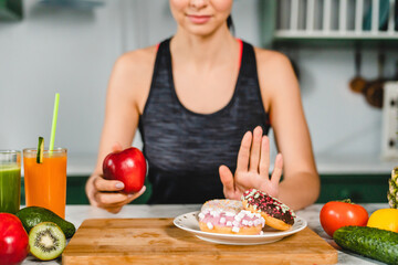 Young fit girl refuses eating doughnuts and opts for healthy food in the kitchen. Difficult choice between unhealthy sweet junk meal and vegetables fruits full of vitamins