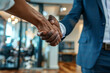 © Emanuel - A welcoming business leader introducing a new team member into the firm, extending a handshake amidst a modern workspace with team members visible behind